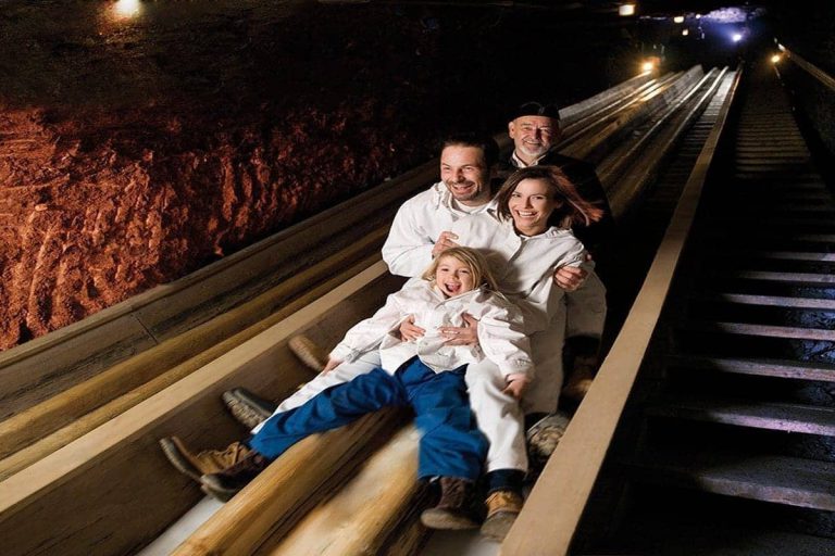 Familie rutscht lachend in Schutzkleidung auf Holzbahn im Salzbergwerk bei Salzburg