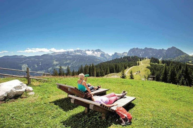 Zwei Menschen entspannen auf Holzliegen mit Blick auf Berge und Almwiese bei Salzburg